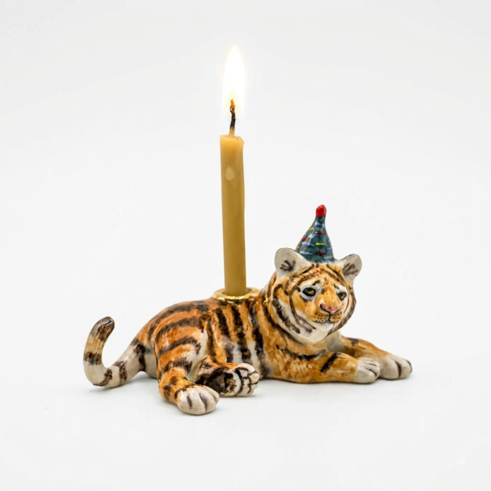 A ceramic tiger figurine candle holder on a white background, featuring a lit candle inserted into its back and a small party hat on its head.