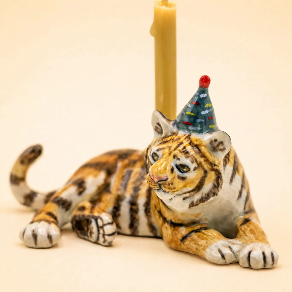 A ceramic tiger figurine lying down, decorated with orange, black, and white stripes, wearing a small blue party hat with a red pom-pom. A yellow candle is placed behind its head on a plain beige background.