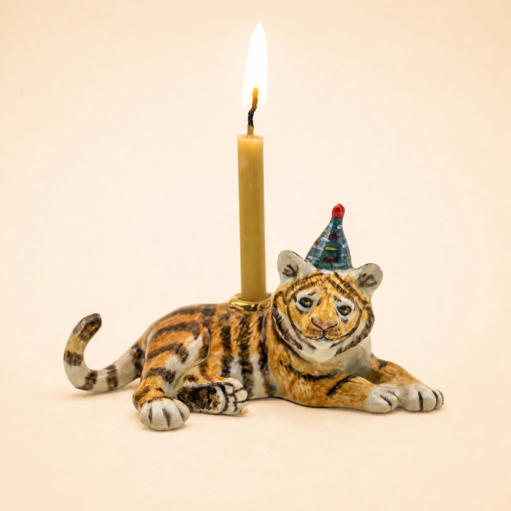 A small tiger figurine candle holder with a lit candle inserted behind its back and a small party hat on its head, photographed against a plain warm beige background.