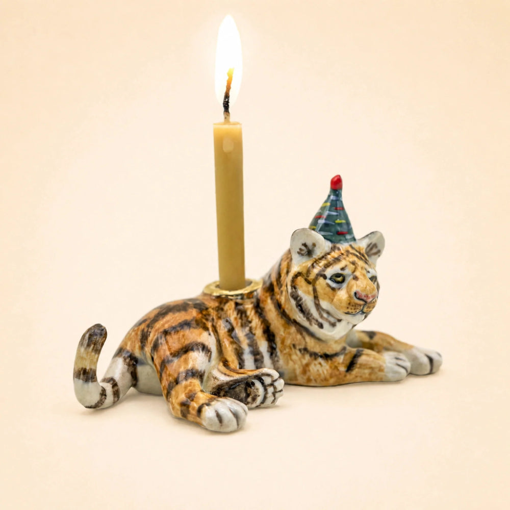 A ceramic tiger figurine candle holder with a lit candle inserted into its back, wearing a small party hat. The figurine is detailed with realistic orange, black, and white tiger stripes and is set against a solid, warm beige background.