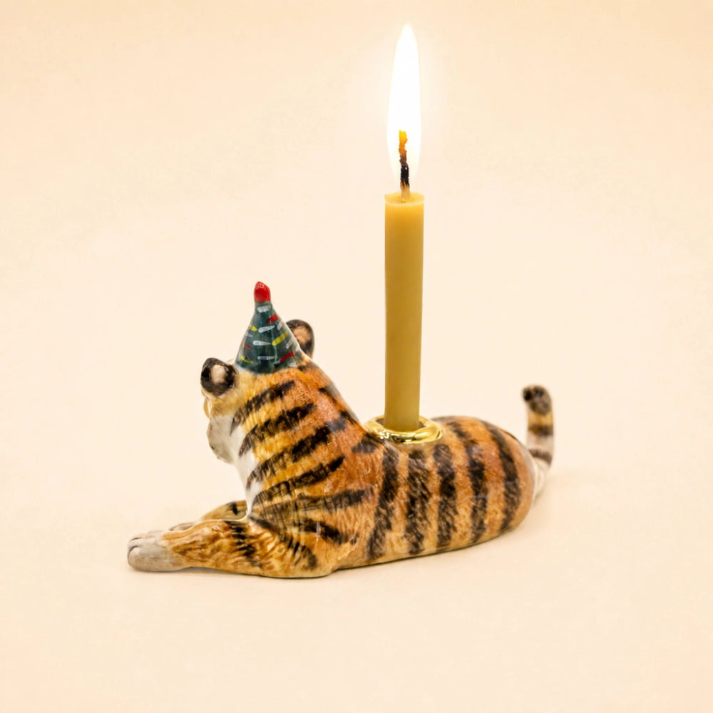 A tiger-shaped candle holder with a lit candle inserted behind it. The tiger is posed lying down and wears a small party hat.