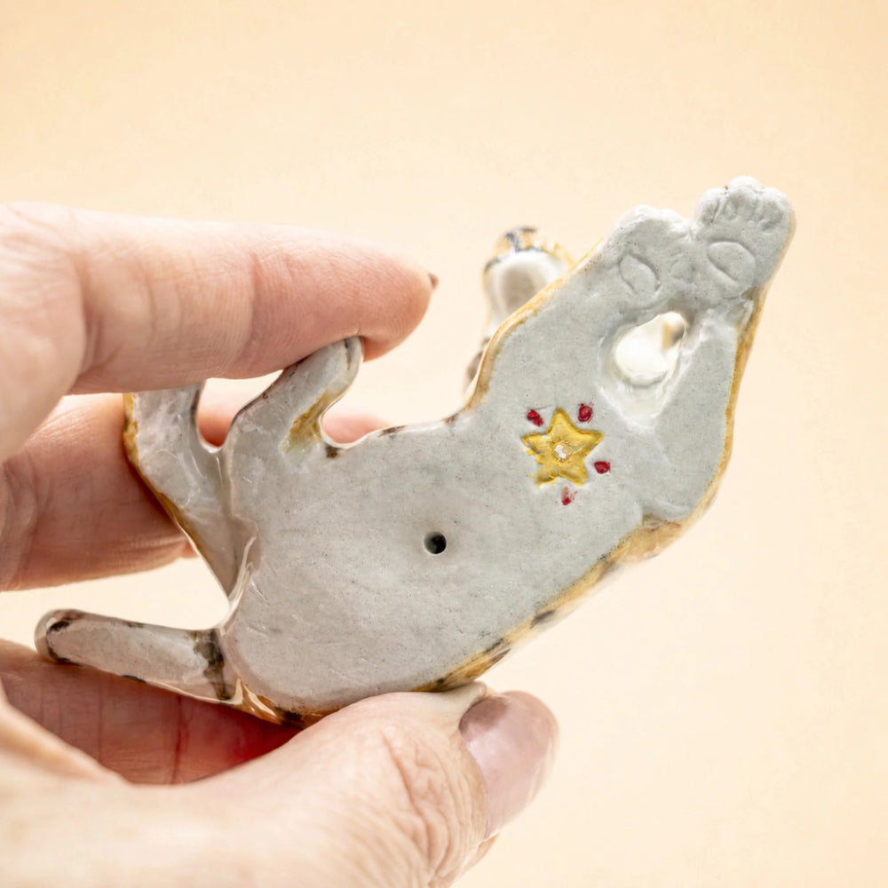 A hand holding a small ceramic figurine of a cat lying on its back, decorated with a yellow star and red dots on a light gray glaze, against a solid beige background.