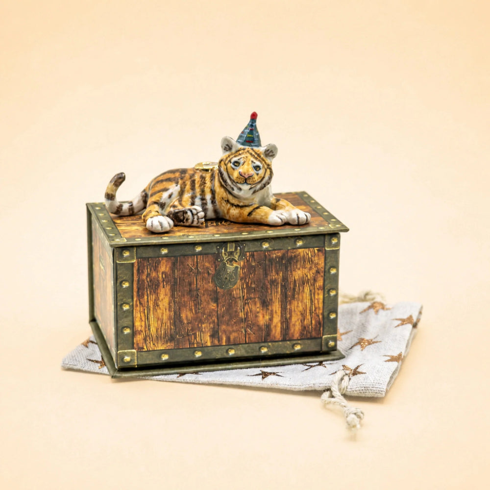 A decorative wooden chest with a small tiger figurine lying on top and wearing a party hat, placed on a light fabric pouch against a warm beige background.