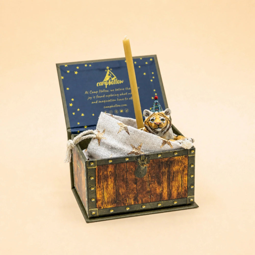 A miniature wooden treasure chest keepsake box with an open lid, a blue starry insert, a tall yellow candle, a small plush tiger toy, and a white drawstring bag on a warm beige background.