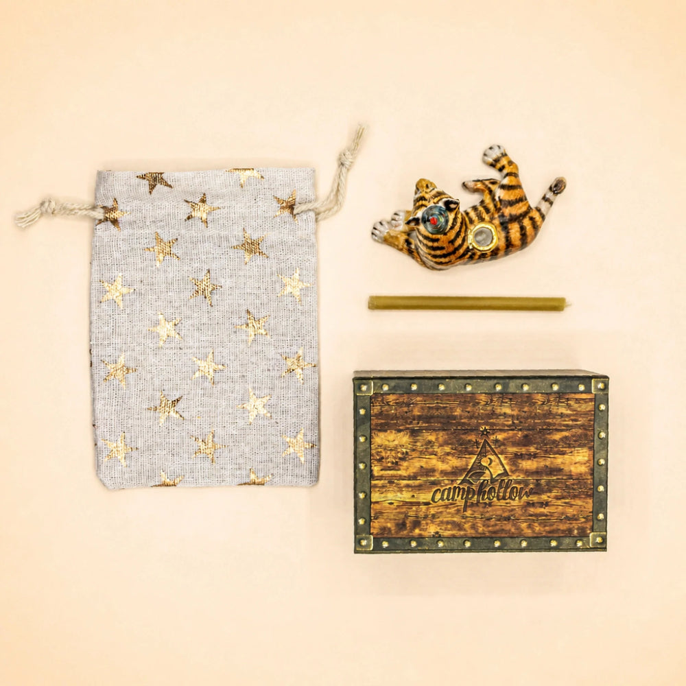A styled flat-lay of a camping gear set on a warm beige background, featuring a light gray drawstring pouch with gold stars, a small tiger-shaped candle holder, a brass rod, and a rustic wooden box with metal studs and the brand name 'camp letter' printed on it.