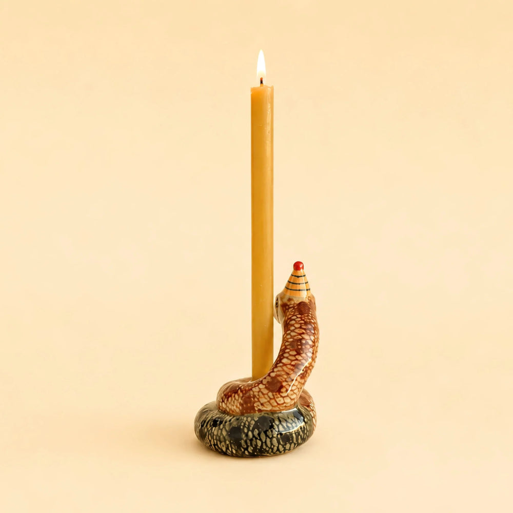 A lit taper candle standing in a sculpted snake holder with a coiled body, patterned scales, and a small red accent on the head, set against a warm beige background.