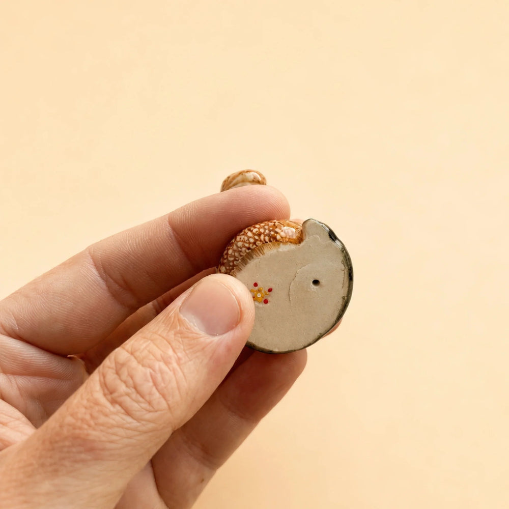 A hand holding a small, hand-painted wooden charm shaped like a hedgehog, set against a solid, warm beige background.