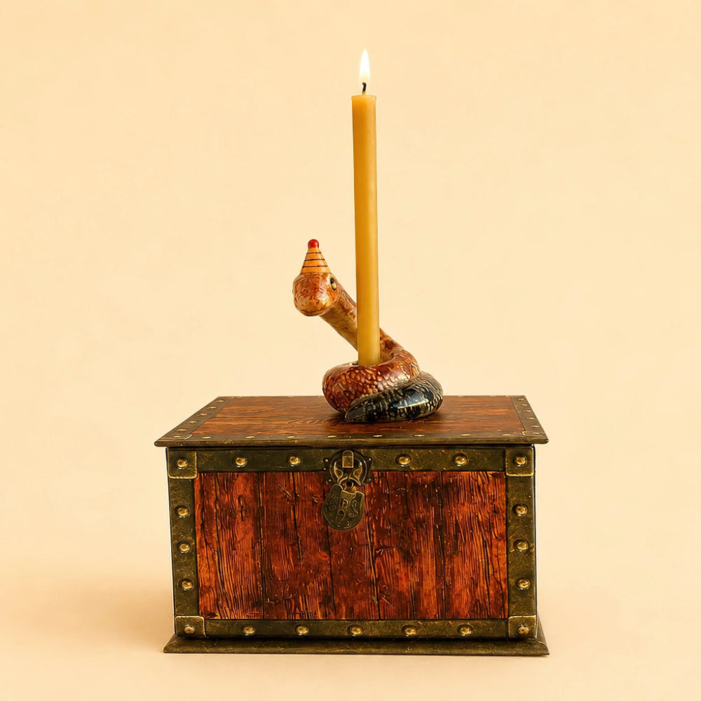 A decorative candle holder shaped like a coiled snake resting on a small wooden treasure chest, with a single lit yellow candle inserted. The chest has brass-toned metal trim and a hanging padlock detail, set against a plain beige background.