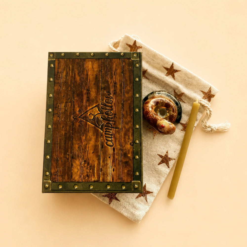 A rustic wooden box with dark green metal trim and brass studs, featuring an embossed logo that reads 'campfire & more', styled on a beige background next to a star-patterned drawstring pouch, a seashell, and a green candlestick.