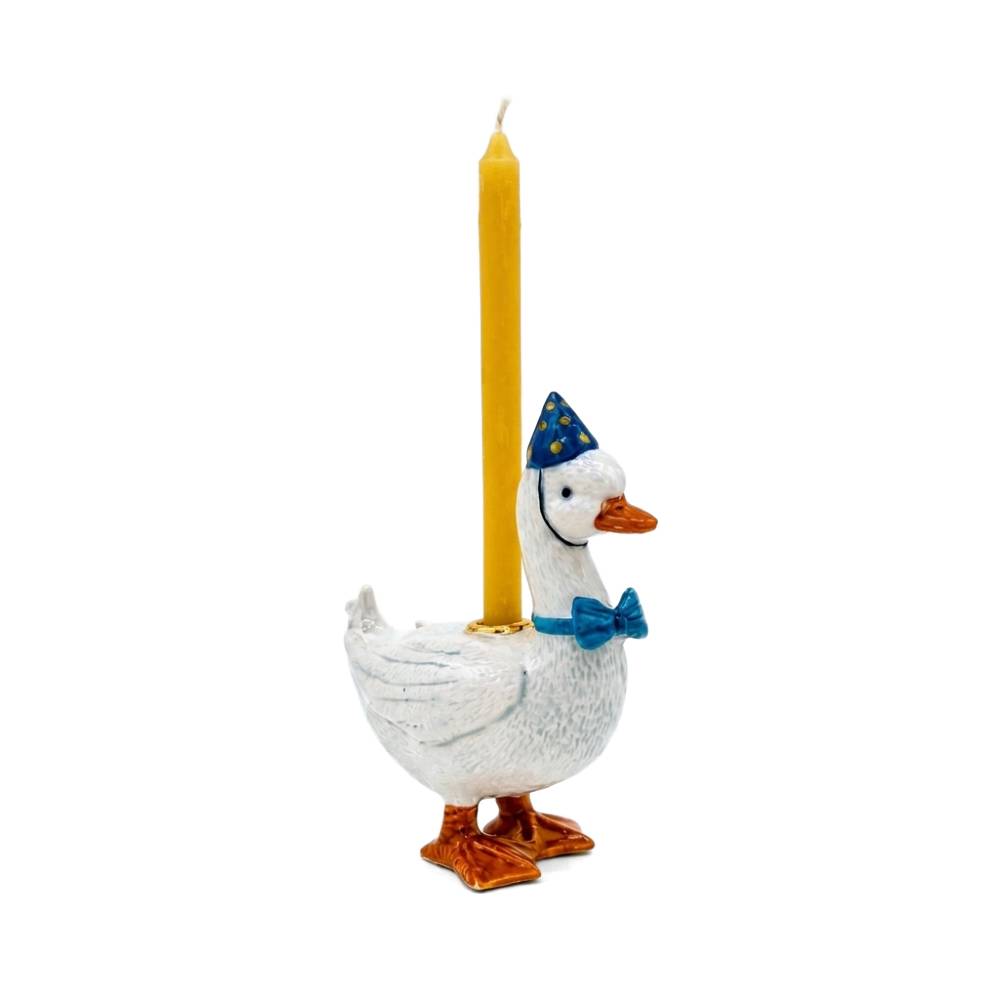 A white goose-shaped candle holder with orange feet and beak, wearing a blue party hat and blue bow tie, holding a single yellow taper candle against a plain white background.