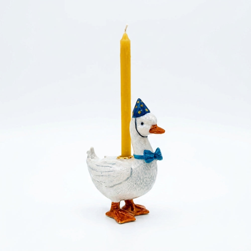 A white goose-shaped candle holder with orange feet and beak, wearing a blue party hat and blue bow tie, holding a single yellow taper candle against a plain white background.