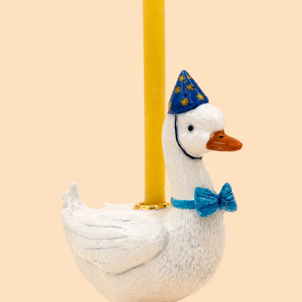 A white goose figurine wearing a blue party hat with yellow stars and a blue bow tie, attached to a yellow pole against a light beige background.