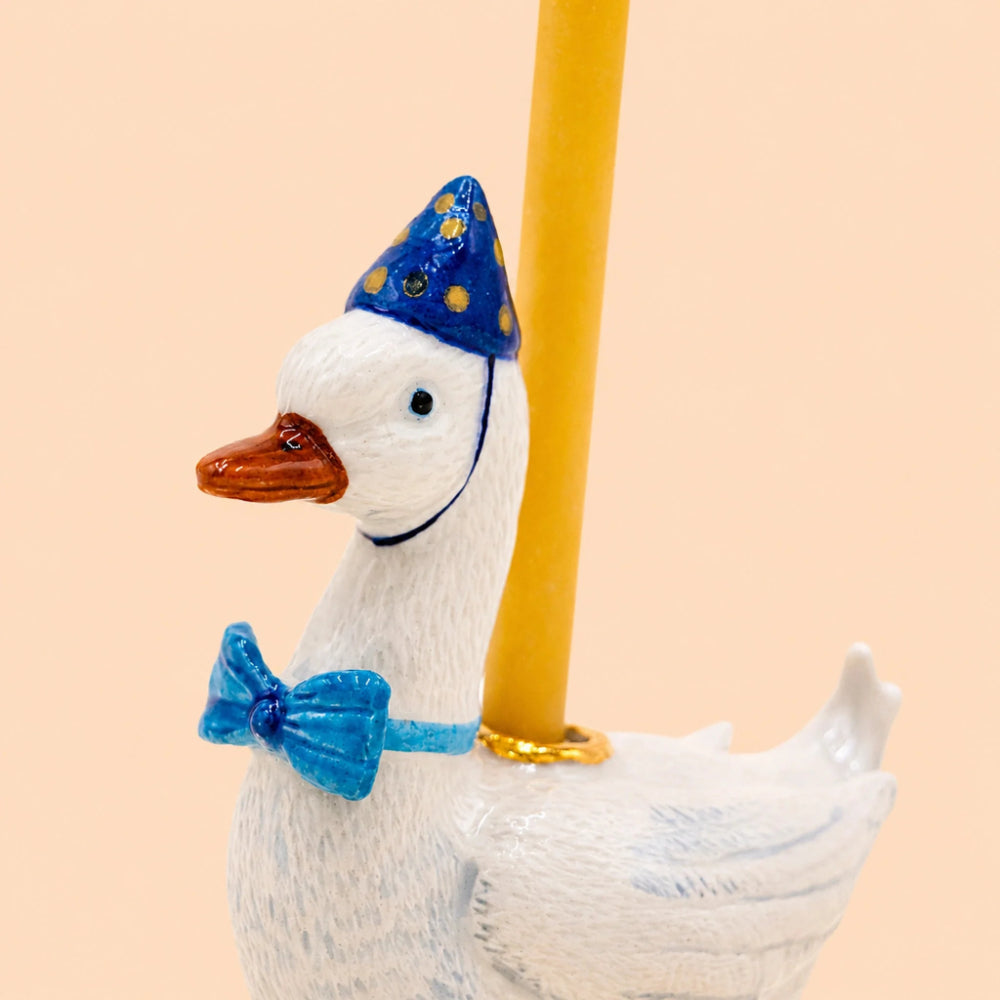 A close-up of a white decorative duck figurine wearing a blue party hat with gold stars and a blue bow tie, with a yellow pole behind it against a soft peach background.
