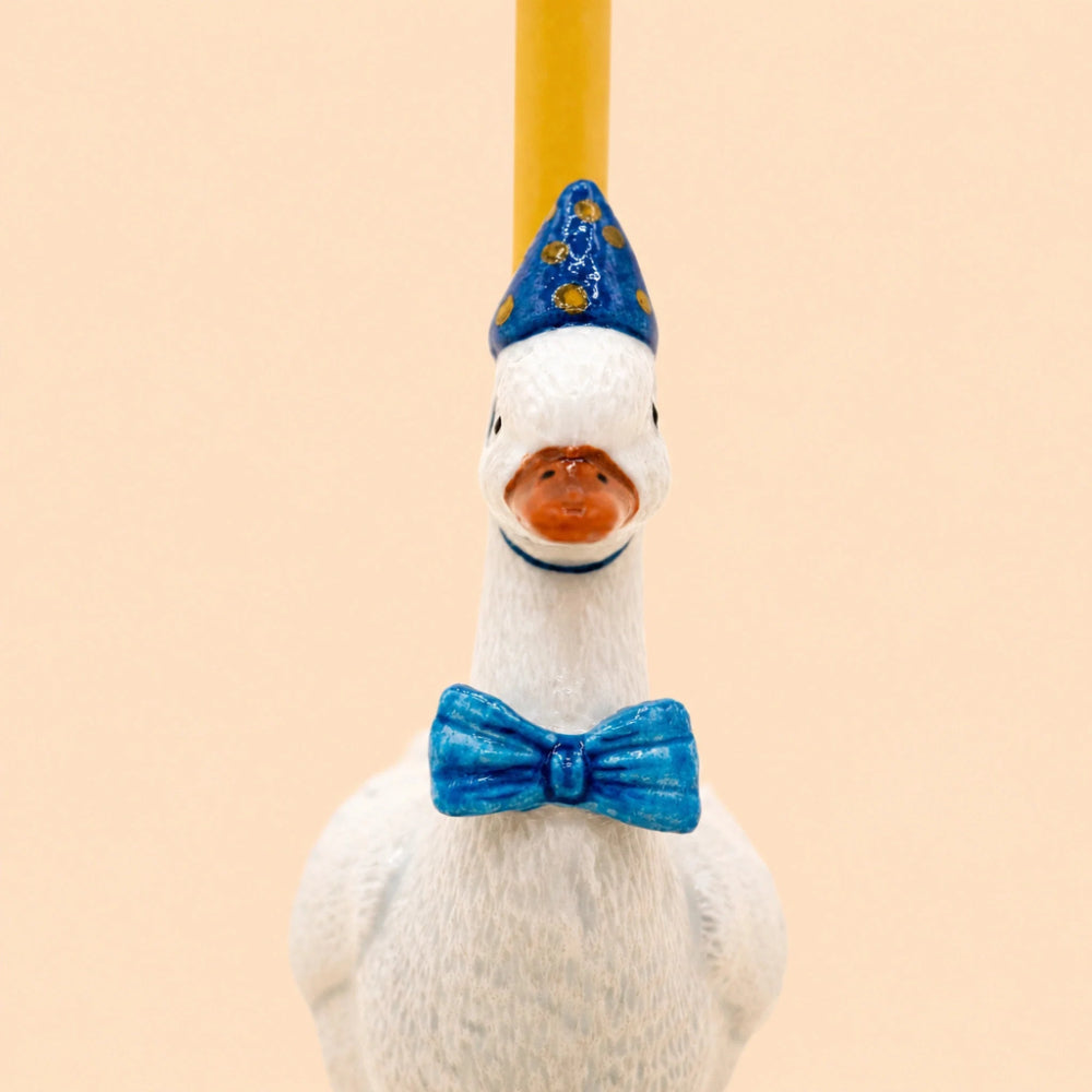A white goose figurine with a blue bow tie and a blue party hat with yellow stars, holding a yellow candlestick or rod upright on its beak, against a solid pale peach background.