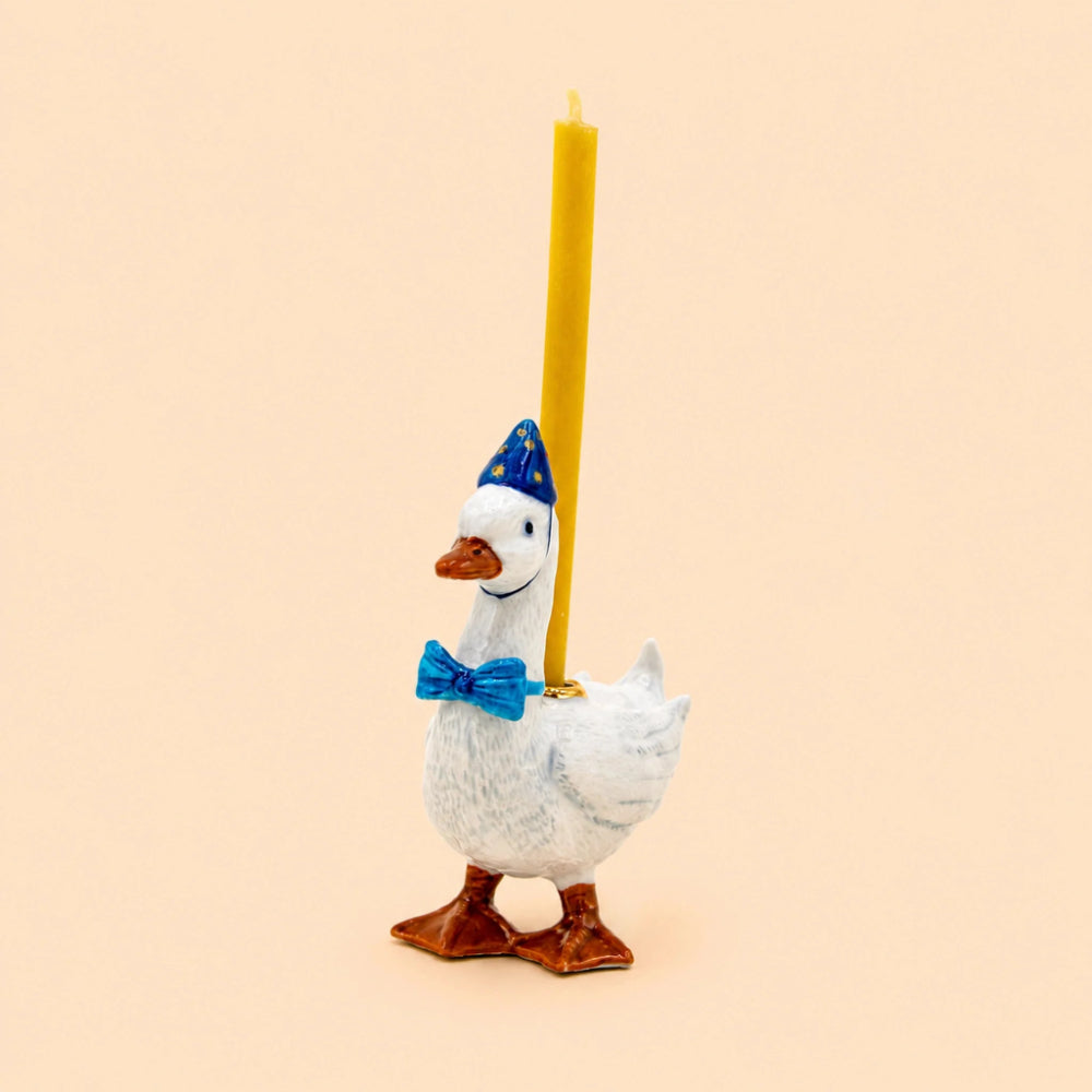 A white goose figurine holding a tall yellow candle, wearing a blue party hat and blue bow tie, set against a solid pale peach background.
