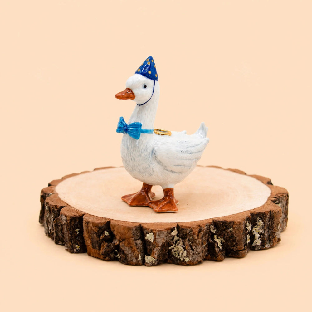 A white decorative duck figurine wearing a blue party hat and blue bow tie, standing on a round natural wood slice against a warm beige background.