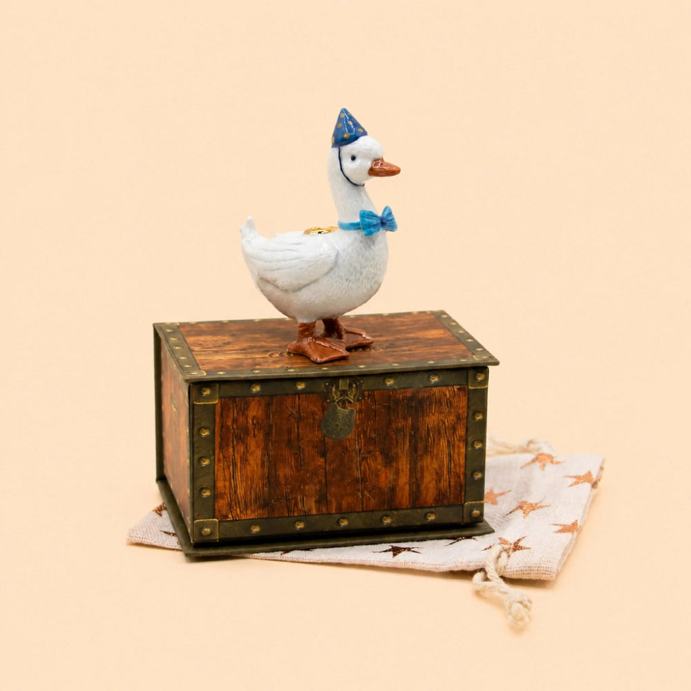 A white goose figurine wearing a blue party hat and blue bow tie, perched on top of a small rustic wooden chest with brass-style corner hardware. The chest sits on a cream drawstring bag with small star motifs, against a plain warm beige background.