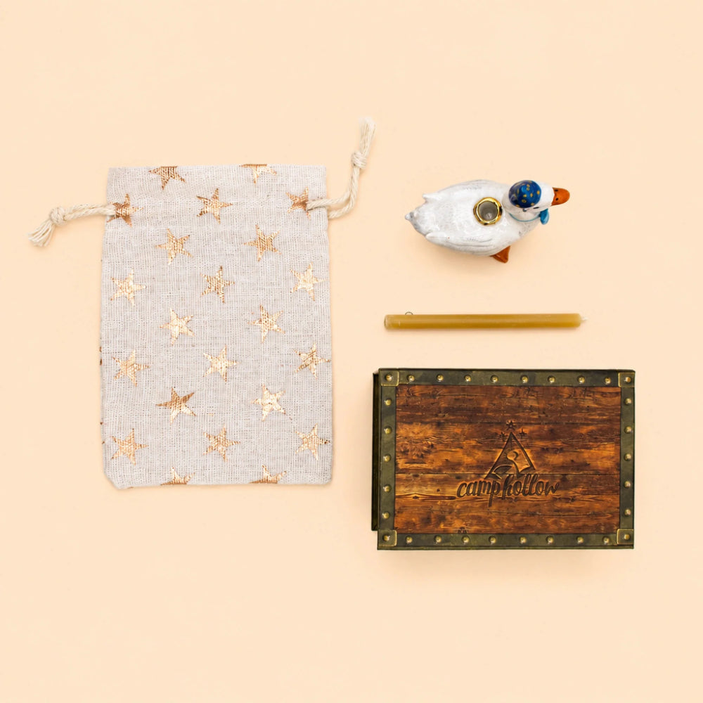 A styled flat-lay of a gift set on a solid pale peach background, featuring a light gray linen drawstring pouch with metallic gold stars, a small white duck figurine with a blue beak, a yellow beeswax candle, and a rustic wooden keepsake box with brass studs and the brand name 'campfire' engraved on the front.