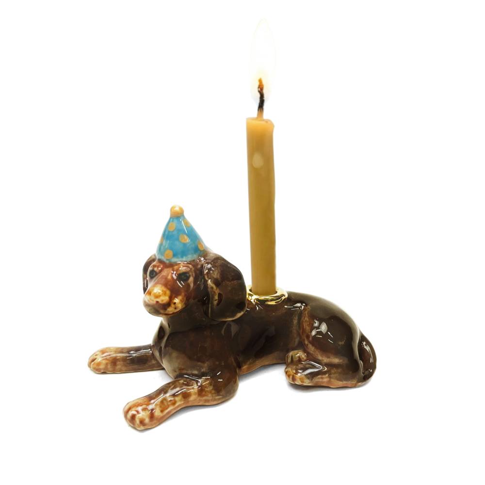 A ceramic dachshund candle holder with a lit candle inserted into its back. The dog is glossy brown with tan spots and wears a blue party hat with gold stars, set against a plain white background.