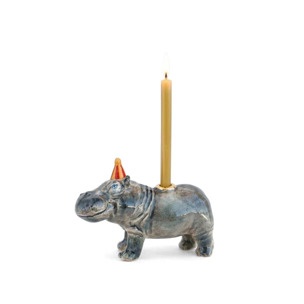 A hippopotamus-shaped candle holder with a blue-gray glazed finish and a small orange party hat, holding a lit gold candle against a plain white background.