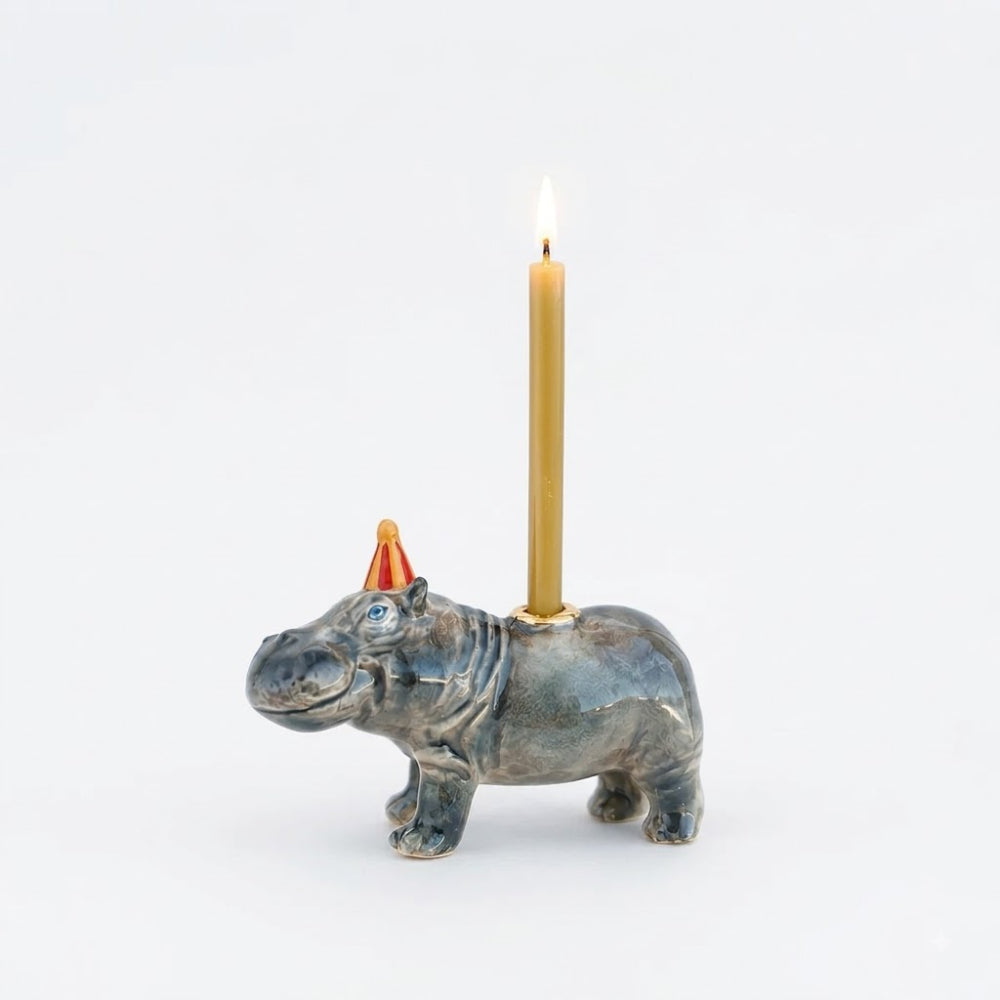 A hippopotamus-shaped candle holder with a blue-gray glazed finish and a small orange party hat, holding a lit gold candle against a plain white background.