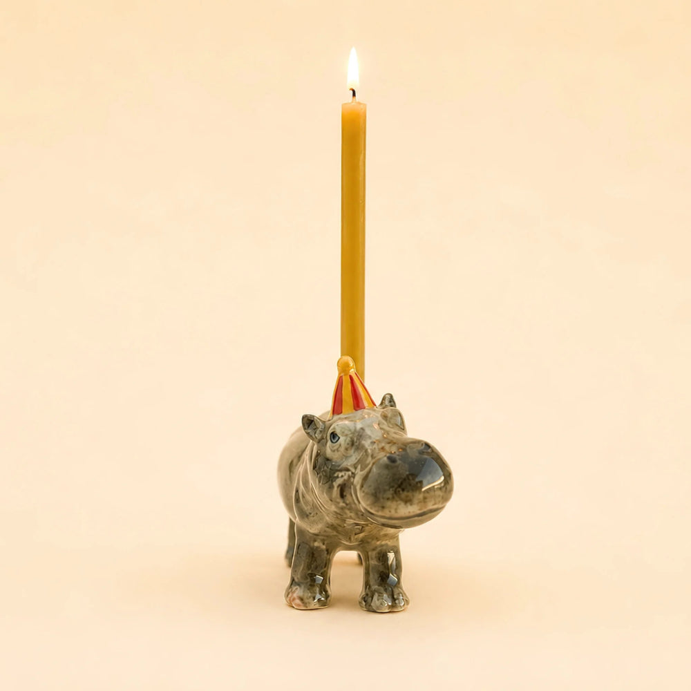 A ceramic hippopotamus candle holder with a lit yellow taper candle inserted behind its head, set against a solid warm beige background.