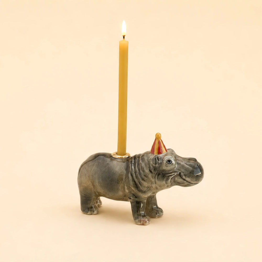 A ceramic hippopotamus candle holder with a lit yellow candle inserted into its back, featuring a small red and yellow party hat on its head, set against a solid beige background.
