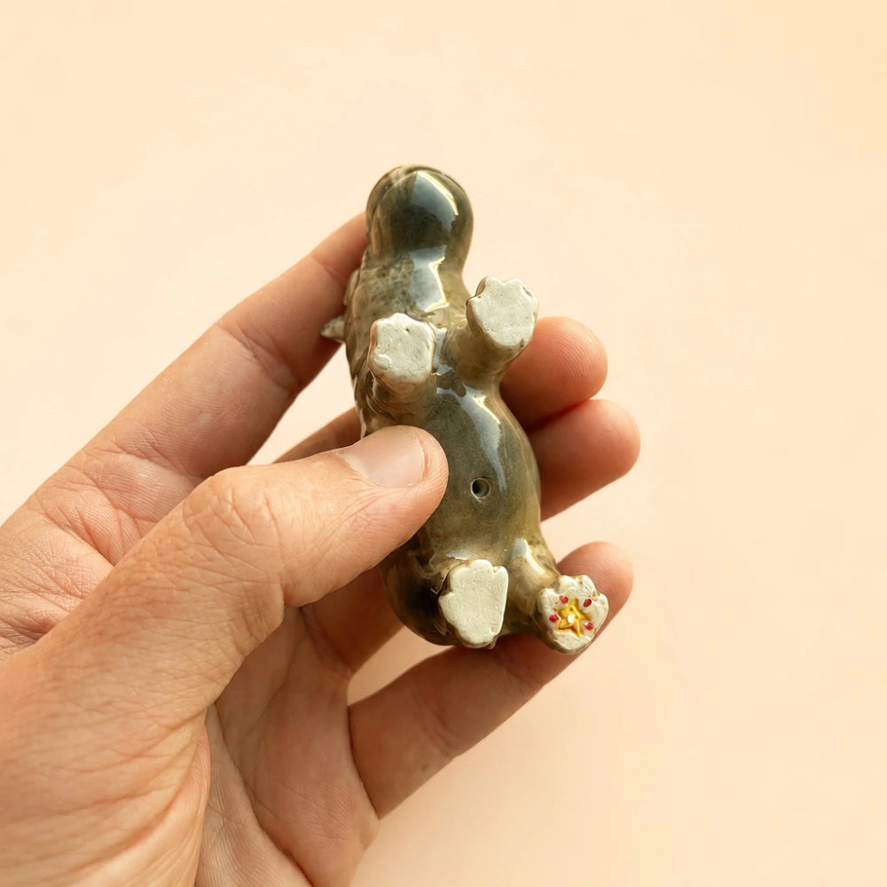A hand holding a small glossy ceramic figurine of a rabbit or hare, shown from the back. The figurine has a dark gray-green glazed body with white painted details on its feet and a small yellow and white floral accent on one paw, set against a solid light beige background.