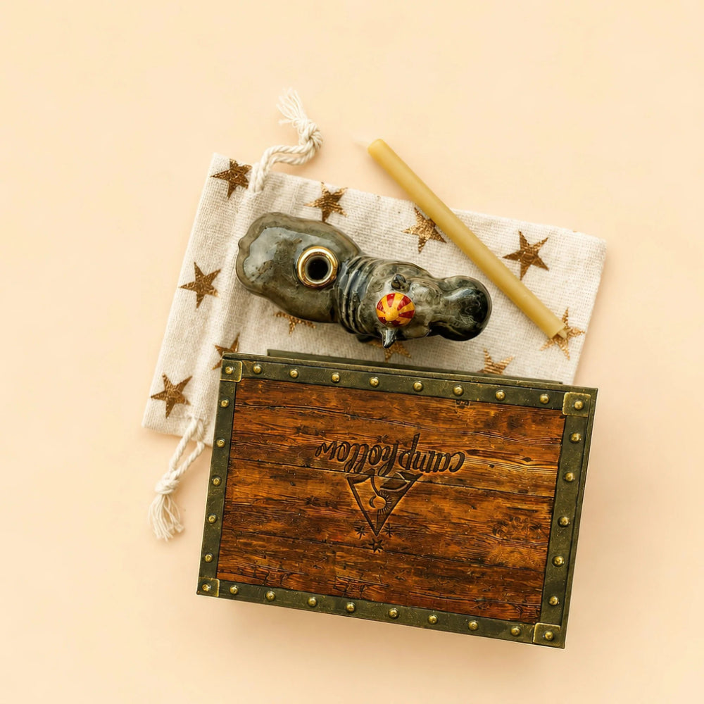 A styled flat-lay featuring a dark brown wooden box with brass studs and a carved 'monopoly' logo, accompanied by a gray ceramic pipe, a yellow taper candle, and a cream fabric pouch decorated with gold stars, all arranged on a solid pale peach background.