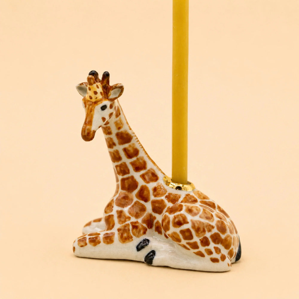A ceramic giraffe candle holder with a white base and brown spots, holding a yellow candle, set against a solid pale beige background.