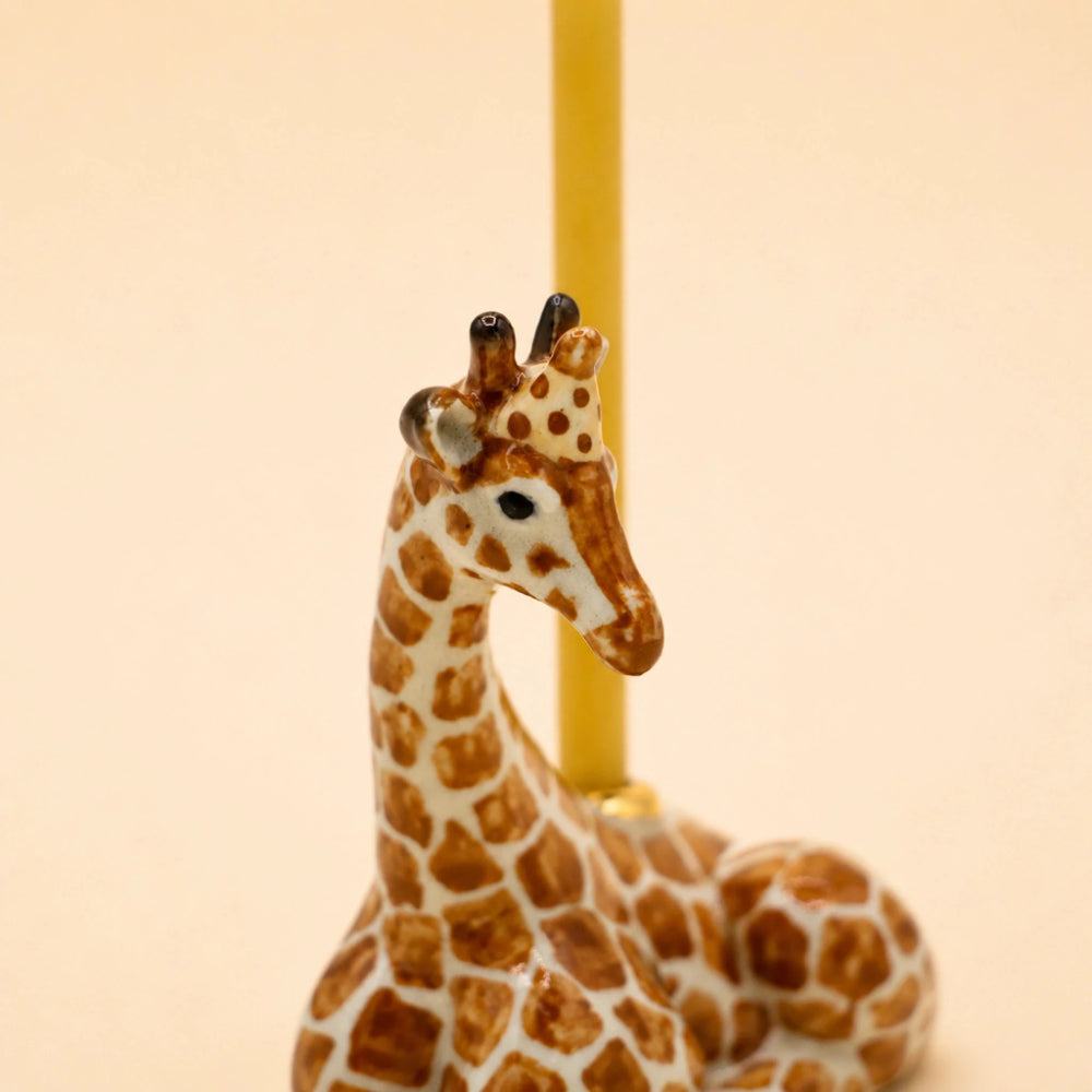 A small giraffe figurine with brown and cream spots, seated in front of a vertical yellow pole on a plain warm beige background.