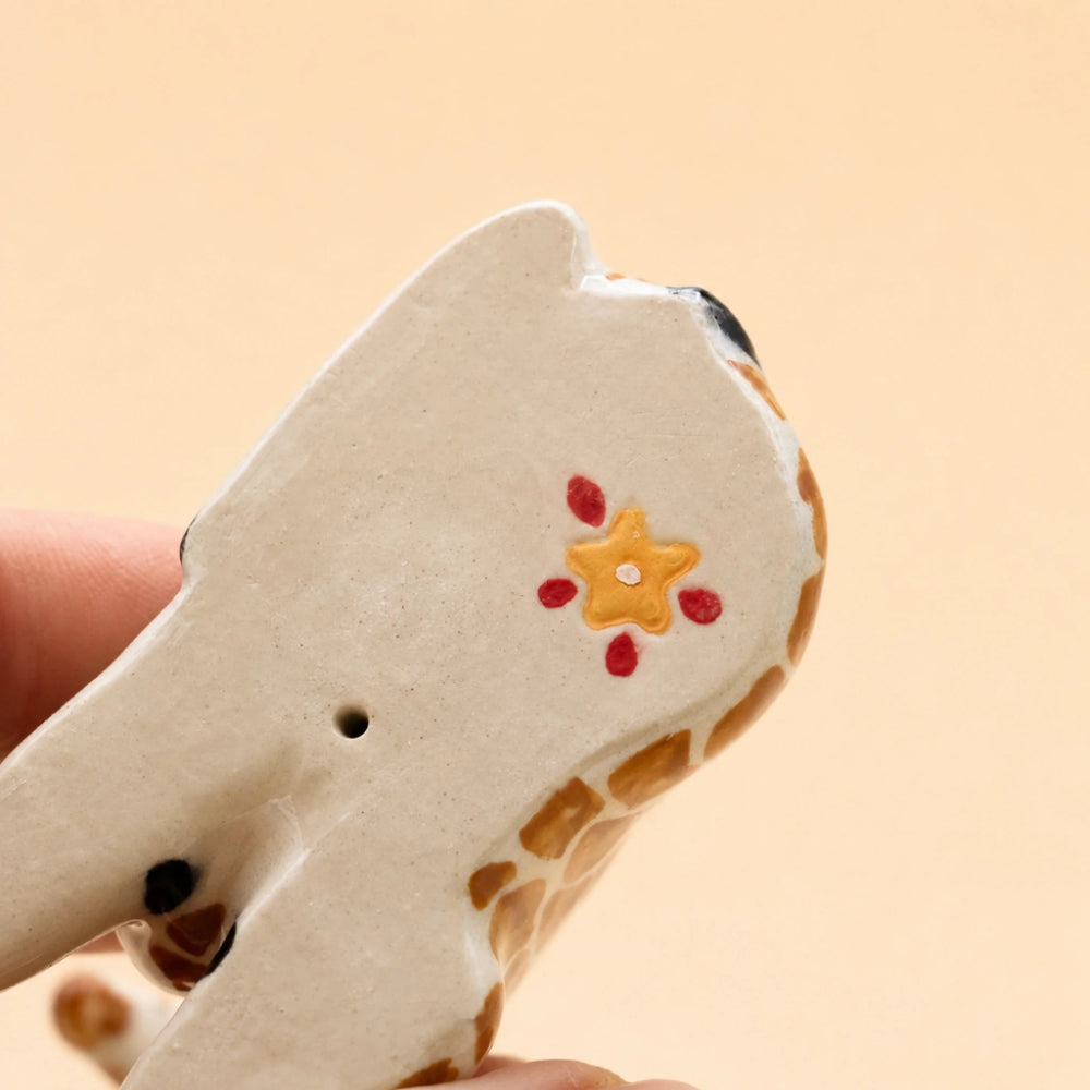 A close-up of a hand holding a small hand-painted ceramic giraffe figurine against a solid beige background.