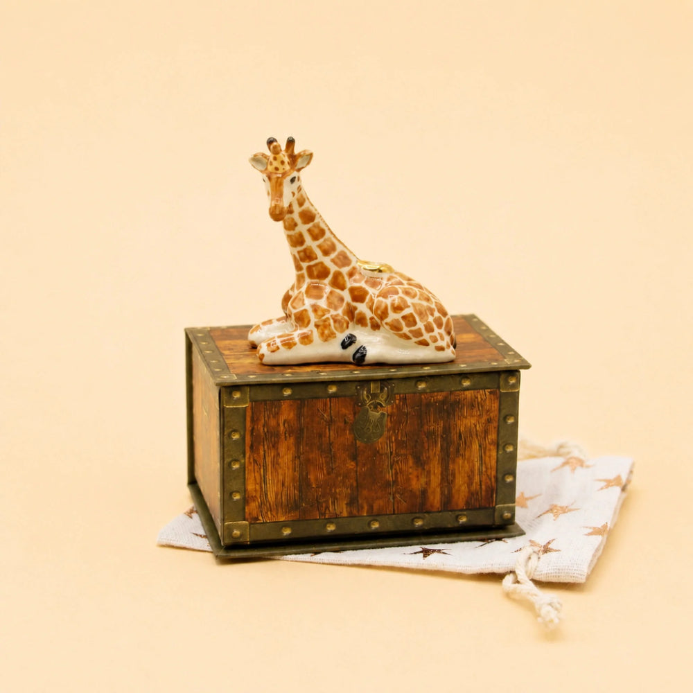 A giraffe figurine resting on top of a small wooden chest with brass-tone corner accents, placed on a white drawstring bag with star prints against a warm beige background.