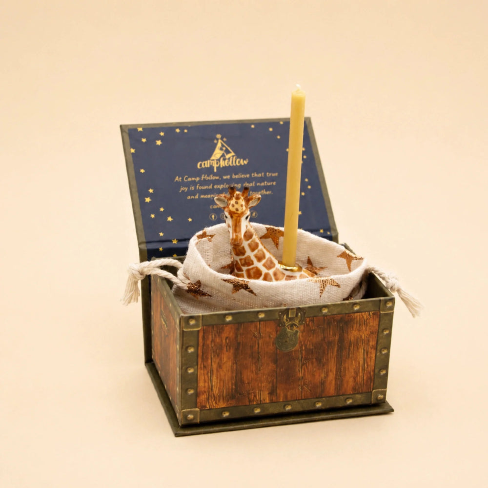 A decorative gift box set with a small wooden treasure chest, an open navy blue lid printed with gold stars and a giraffe illustration, a white fabric pouch with a giraffe print, and a single yellow candle standing upright inside.
