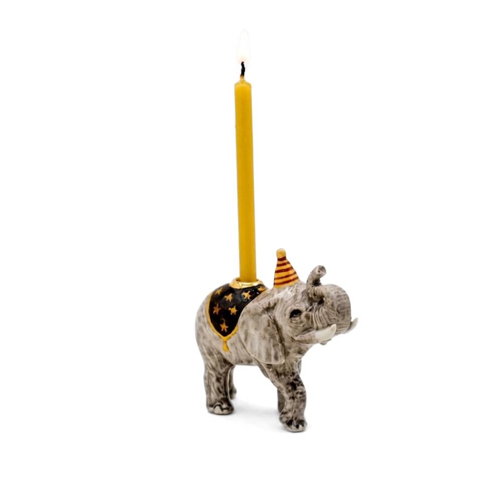 A ceramic elephant candle holder with a gray and white marbled pattern, featuring a black and gold star-patterned saddle and a small orange-and-yellow striped party hat. A single yellow candle is inserted into the back of the holder, with a small flame lit, set against a plain white background.
