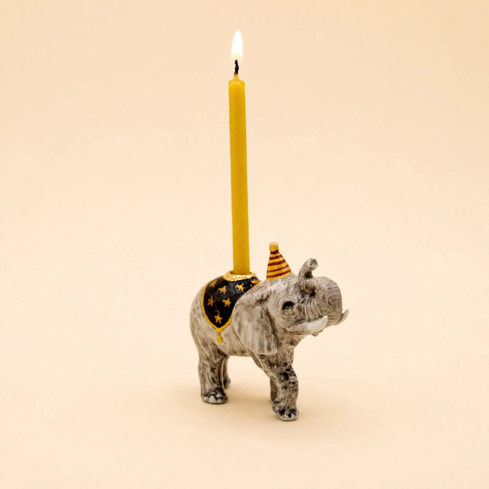 A small elephant-shaped candle holder in gray and white marbled ceramic, holding a tall yellow candle with a black star-patterned saddle and a small striped party hat, on a plain beige background.