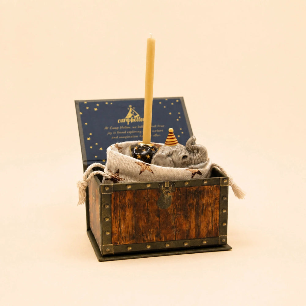 A miniature Christmas jingle bell gift set in a small wooden chest. The set includes a blue starry lid with gold lettering, a tall cream candle, a gray fabric sack, and a small toy jingle bell with a striped cone topper, all on a solid cream background.