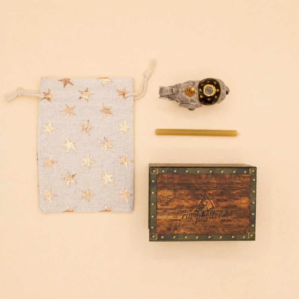 A gift set laid out on a warm beige background, including a light blue drawstring pouch with metallic gold star prints, a small gray decorative object with a gold center, a thin gold rod, and a rectangular wooden box with metal corner studs and the word 'campfire' on the front.