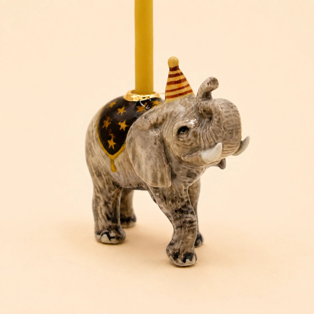 A small elephant figurine with a gray and black spotted body, white tusks, a black saddle with gold stars, a gold trim, and a striped orange and yellow party hat. A yellow candle is inserted into the top of the figurine, set against a solid pale peach background.