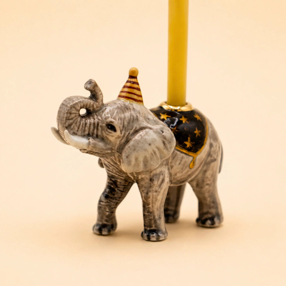 A small elephant figurine holding a yellow candle in its trunk, with a black and gold star-patterned saddle and a striped party hat, photographed against a plain beige background.