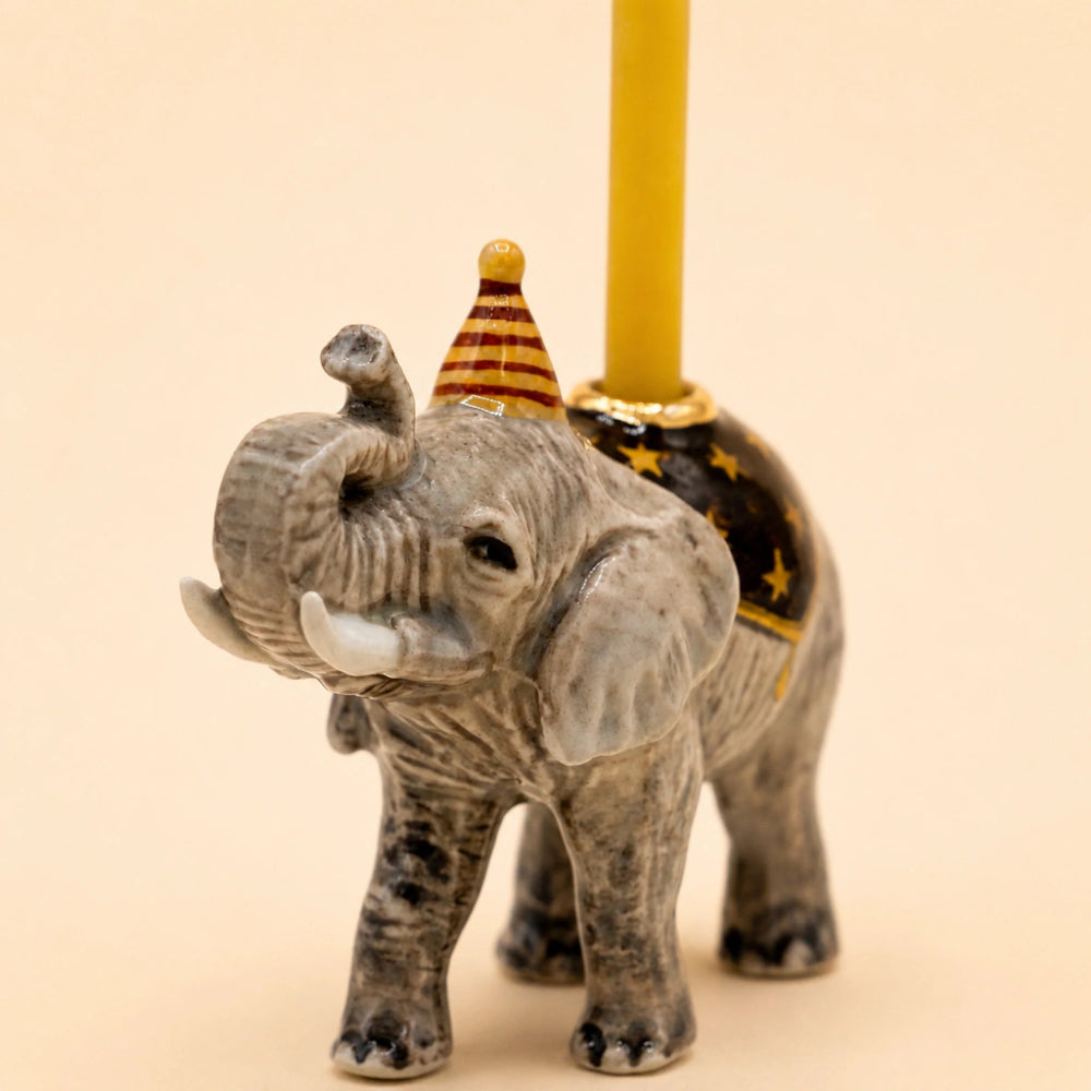 A small gray elephant figurine with a party hat and a yellow candle holder inserted behind it, set against a plain beige background.