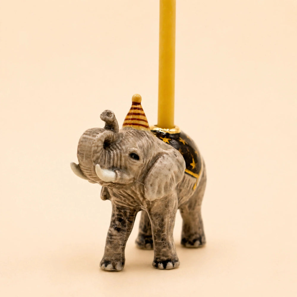 A small elephant figurine holding a yellow pole, decorated with a striped party hat and a black-and-gold saddle, on a plain beige background.