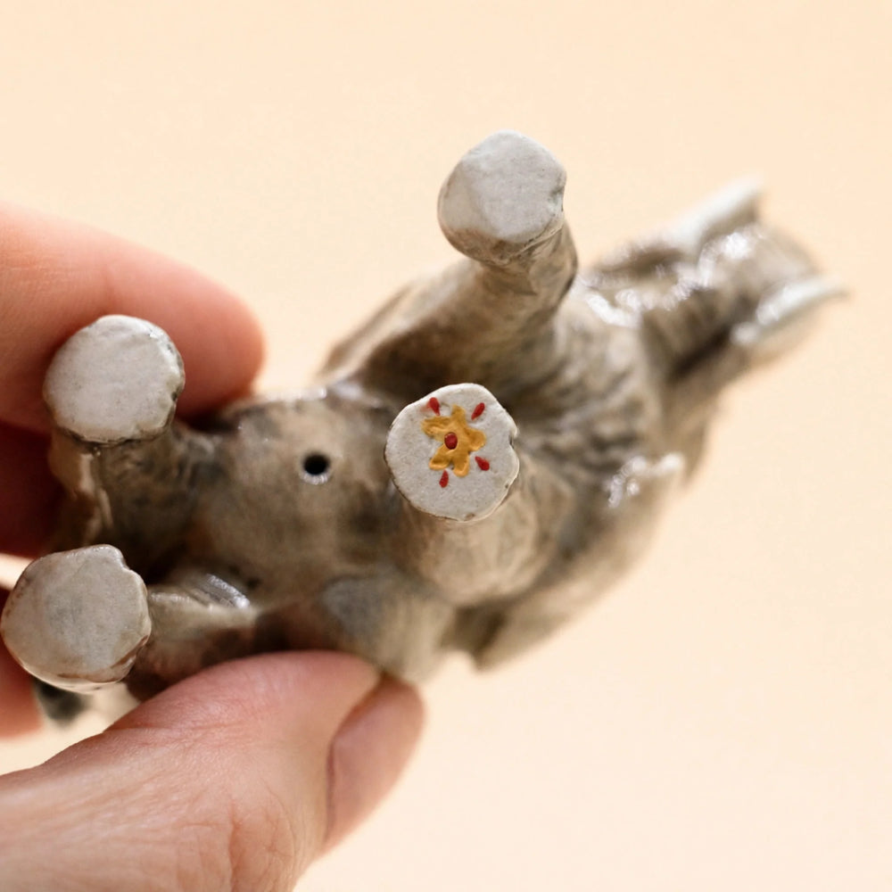 A small gray ceramic figurine of an elephant lying on its back, held in a person's hand against a soft beige background. The figurine has a small painted yellow and red flower detail on one of its feet.