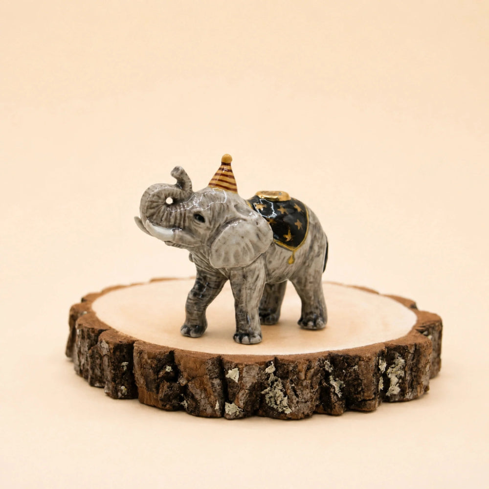 A small gray elephant figurine wearing a striped party hat and a black-and-gold saddle, standing on a round natural wood slice against a warm beige background.