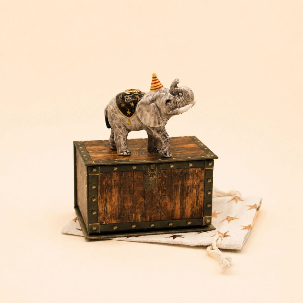 A small gray elephant figurine wearing a gold saddle and a party hat, standing on top of a rustic brown wooden chest with metal corner accents. The chest sits on a white drawstring pouch with gold star prints against a warm cream background.