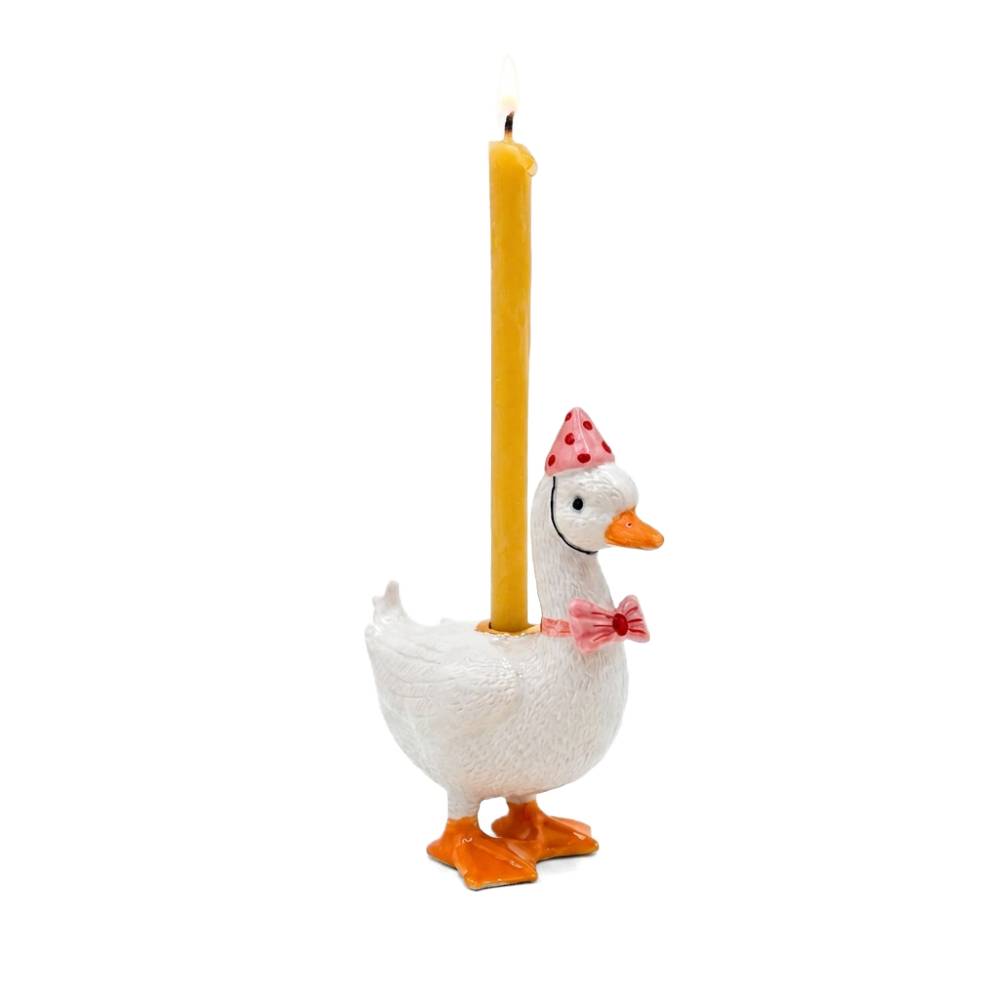 A white goose-shaped candle holder with orange feet and beak, wearing a pink party hat and bow tie, holding a tall yellow candle that is lit, on a white background.