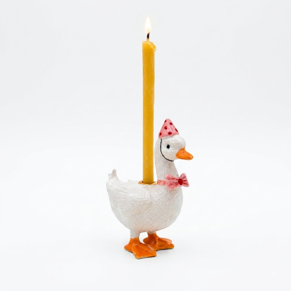 A white goose-shaped candle holder with orange feet and beak, wearing a pink party hat and bow tie, holding a tall yellow candle that is lit, on a white background.