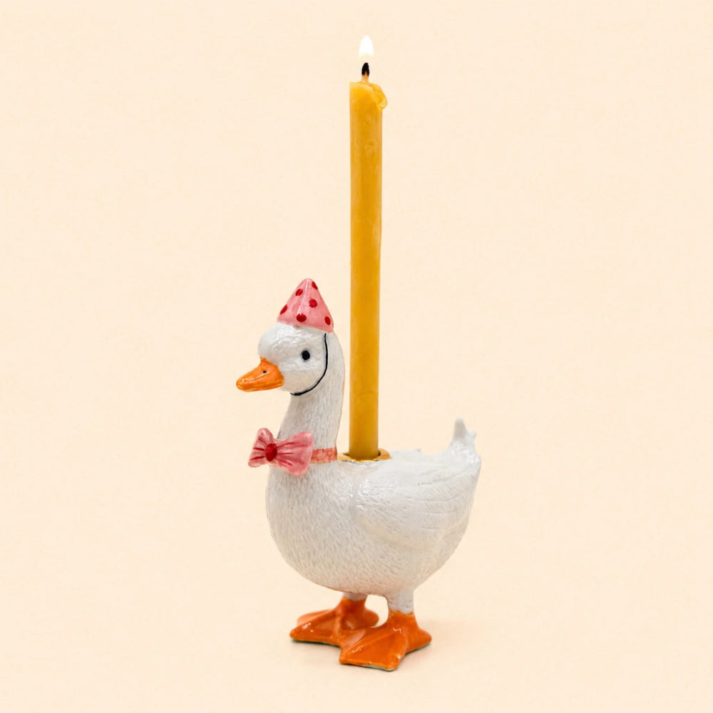 A white duck-shaped candle holder with orange feet and beak, wearing a pink polka-dot party hat and pink bow tie, holding a lit yellow candle against a warm beige background.