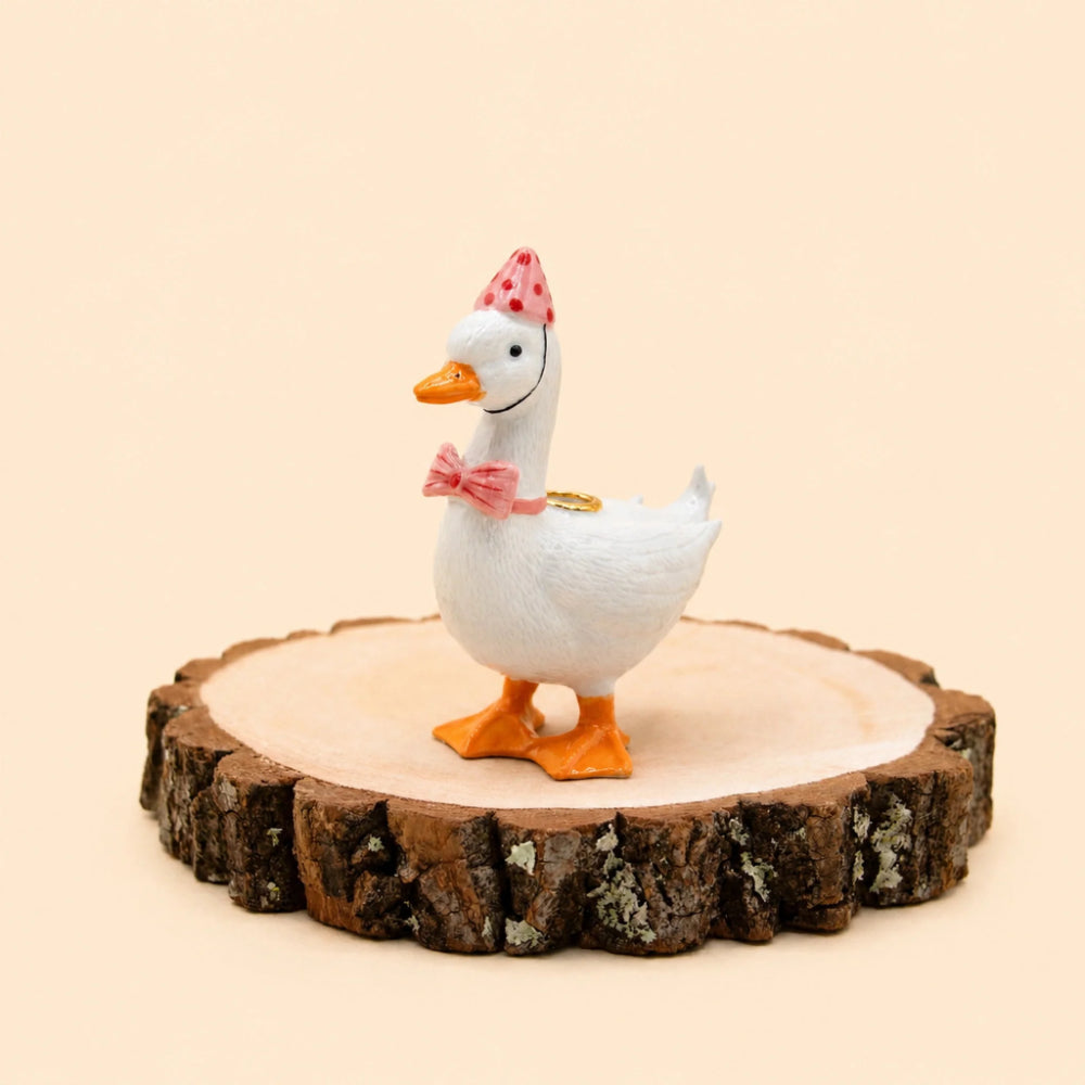 A white decorative duck figurine with orange feet and beak, wearing a pink polka-dot party hat and pink bow, standing on a round natural wood slice against a warm beige background.