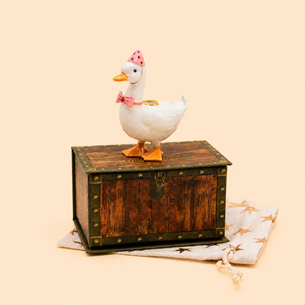 A white duck figurine with orange feet and beak, wearing a pink polka-dot hat and pink bow, standing on top of a rustic brown wooden chest with metal trim. A cream drawstring bag with star prints rests underneath on a solid light beige background.